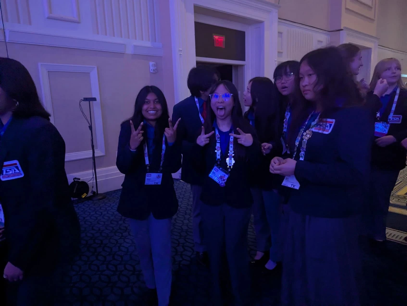 SLHS TSA members in navy competition blazers throwing peace signs in a blue-lit ballroom foyer at the 2026 National TSA Conference